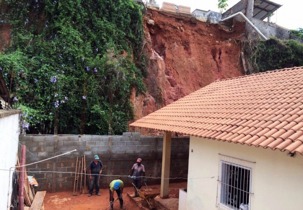 Acima vê-se o barranco que desmoronou e parte dos túmulos do cemitério. Na casa atingida, homens fazem a limpeza